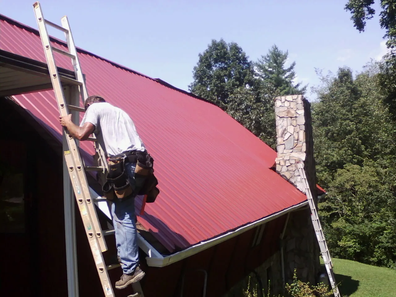 Roofer on a ladder beside a red metal roof and stone chimney — Soffit & Fascia Repair by Roofing Brothers in Cross Fork